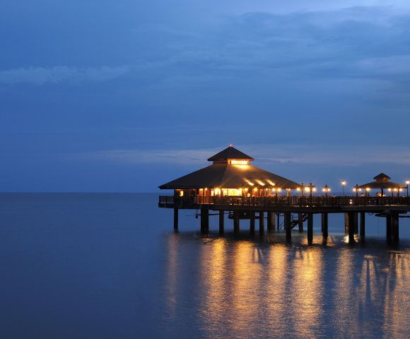 A serene evening scene of a warmly lit wooden pavilion on stilts over calm water. The structure reflects on the ocean, under a tranquil, deep blue sky.