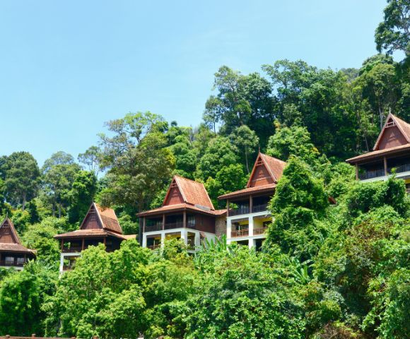 Traditional wooden houses with peaked roofs nestled on a lush, green hillside surrounded by dense tropical trees under a clear blue sky.