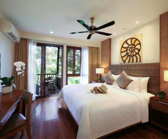 Elegant hotel room with a large bed, three cushions, and a hat. Wooden floor, ceiling fan, and art on the wall. Balcony view of lush greenery.