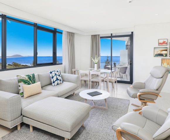 Bright, modern living room with ocean views through large windows. Gray sectional sofa, reclining chair, dining table, neutral tones, and coastal decor.