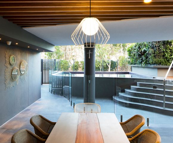 Spacious modern patio with a wooden table, wicker chairs, a stylish white lamp, and a view of a raised pool and garden, creating a serene ambiance.