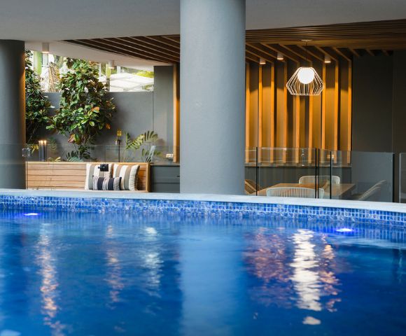 Indoor pool area with blue water, surrounded by tall pillars and modern decor. Wooden paneling, plants, and cozy seating create a relaxing atmosphere.