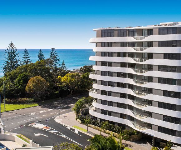A modern, curved apartment building overlooks a scenic coastal road. The ocean and clear blue sky form a serene backdrop, with lush trees adding greenery.