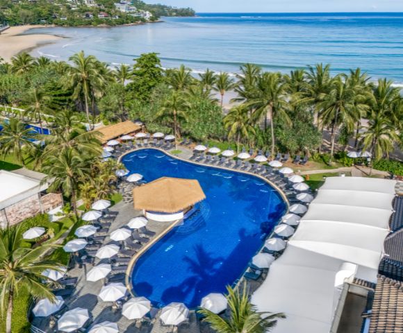 Aerial view of a tropical resort with a lagoon-shaped pool surrounded by umbrellas, palm trees, and a sandy beach next to a calm, blue ocean.