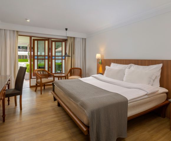 A cozy hotel room with a wooden double bed, white linens, and a gray throw. Natural light streams through a window near a seating area with chairs.