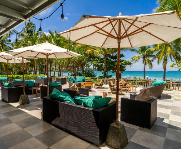 Outdoor seating area with wicker sofas and turquoise cushions under large white umbrellas. Palm trees and ocean view create a tropical, relaxing atmosphere.