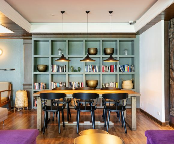 Modern indoor seating area with a wooden table, four black chairs, and overhead pendant lights. A bookshelf with books and vases adds a cozy touch.
