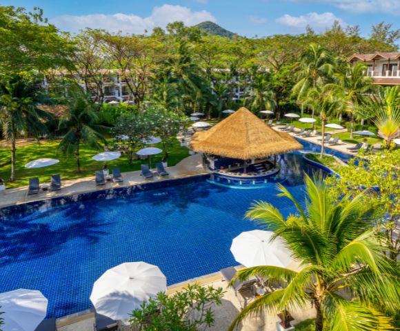 A serene resort pool with deep blue tiles surrounded by palm trees and greenery. White umbrellas and a thatched-roof bar create a tropical vibe.