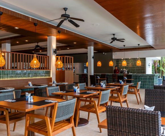 Modern restaurant interior with wooden tables, wicker chairs, and warm lighting. Ceiling fans and hanging lamps create a cozy, inviting ambiance.