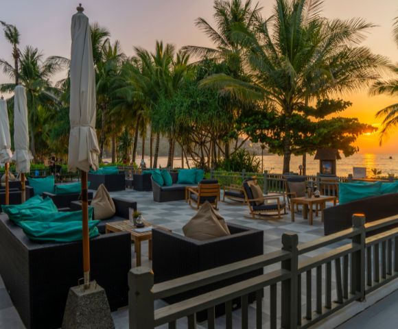 Outdoor lounge with wicker furniture and teal cushions, surrounded by palm trees. A tranquil ocean view at sunset creates a relaxed, tropical ambiance.