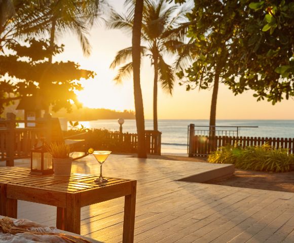A sunset scene at a beachside patio with palm trees. A cocktail glass rests on a wooden table, radiating warmth and relaxation in golden sunlight.