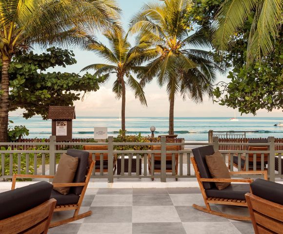 A tranquil beach scene with palm trees framing a view of the ocean. Wooden rocking chairs sit on a tiled patio, offering a serene and inviting atmosphere.