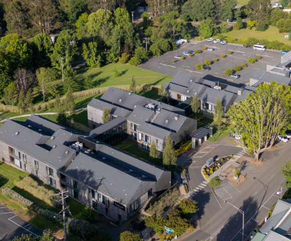 Aerial view of a modern building complex surrounded by lush greenery. The scene is peaceful, with empty parking spaces and tree-lined paths.