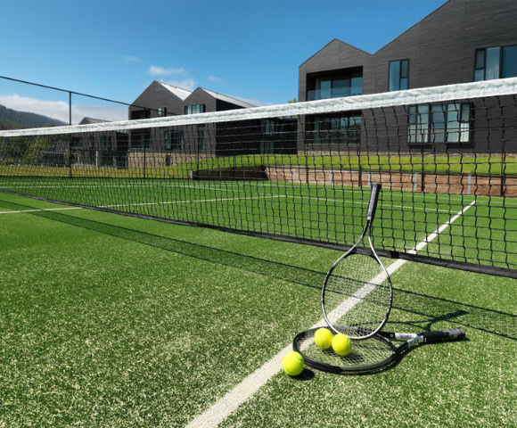 Tennis court with vivid green grass, two rackets, and three yellow balls near the net. Modern black buildings and clear blue sky in the background.