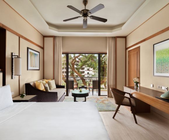 A serene hotel room with a large bed, wicker sofa, desk, and chairs. A balcony overlooks lush greenery, creating a tranquil, inviting atmosphere.