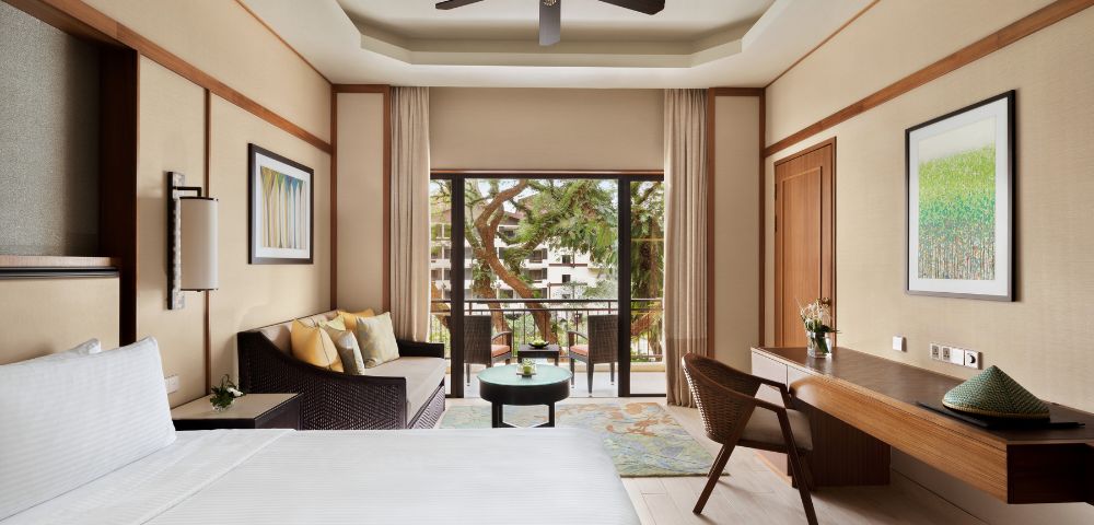 A serene hotel room with a large bed, wicker sofa, desk, and chairs. A balcony overlooks lush greenery, creating a tranquil, inviting atmosphere.