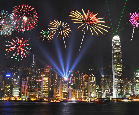 A vibrant fireworks display lights up a city skyline at night, with colorful bursts and beams illuminating tall skyscrapers, creating a festive atmosphere.