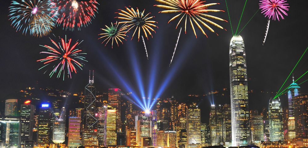 A vibrant fireworks display lights up a city skyline at night, with colorful bursts and beams illuminating tall skyscrapers, creating a festive atmosphere.