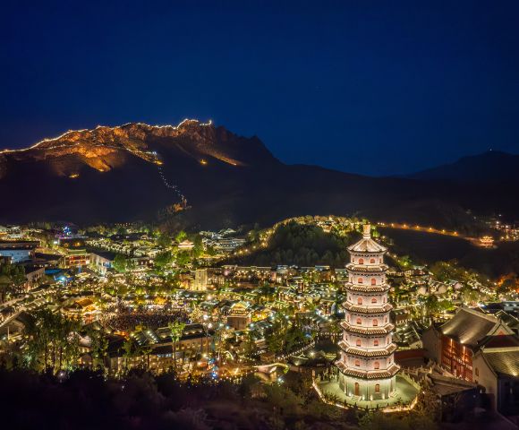 Illuminated mountain town at night with a pagoda in the foreground, glowing buildings, and mountains in the background. The atmosphere is serene and picturesque.