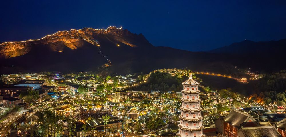 Illuminated mountain town at night with a pagoda in the foreground, glowing buildings, and mountains in the background. The atmosphere is serene and picturesque.
