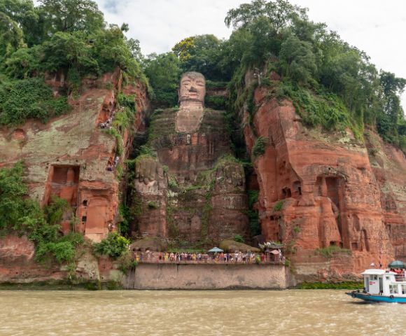 A large stone Buddha statue carved into a lush, green cliffside, overlooking a river with a small tour boat passing by. The scene conveys tranquility.