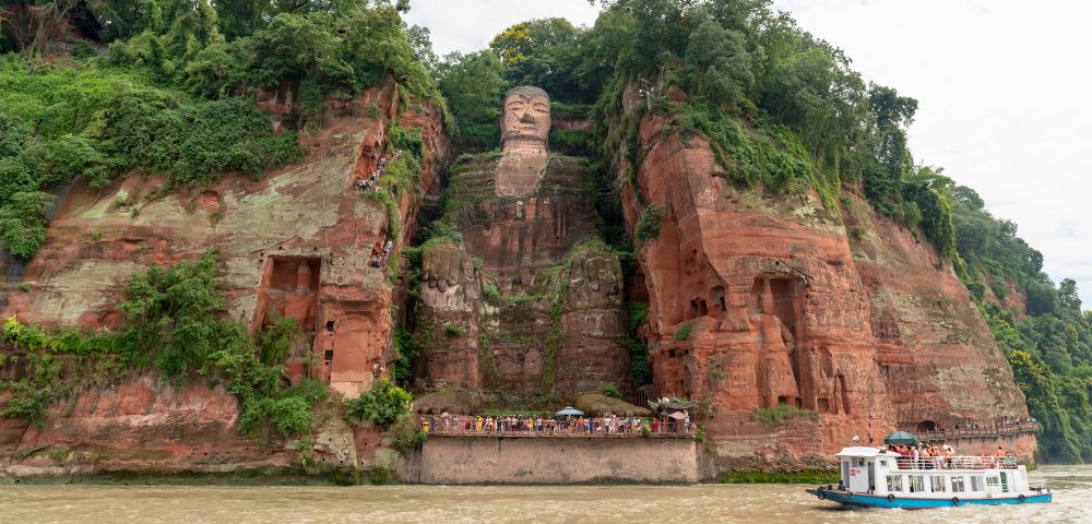 A large stone Buddha statue carved into a lush, green cliffside, overlooking a river with a small tour boat passing by. The scene conveys tranquility.