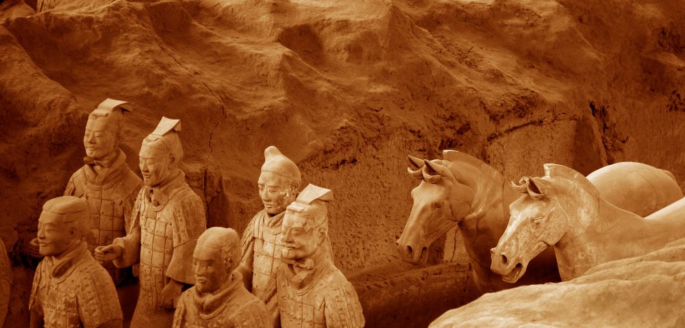 Ancient Terracotta Army sculptures in an excavation pit; lifelike clay warriors and horses, aligned in rows. The scene conveys historical depth and solemnity.