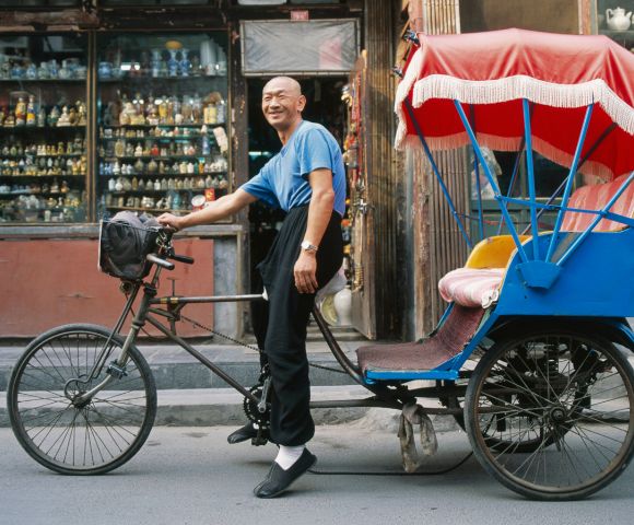 A man in a blue shirt smiles beside a vibrant blue and red rickshaw on a street. Behind him is a shop filled with colorful figurines, creating a lively scene.