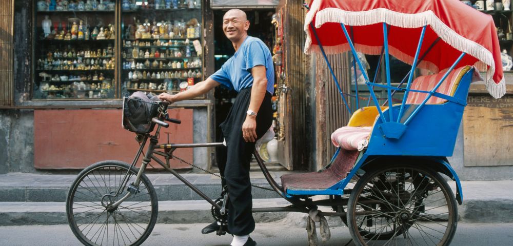 A man in a blue shirt smiles beside a vibrant blue and red rickshaw on a street. Behind him is a shop filled with colorful figurines, creating a lively scene.