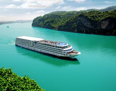 A large white cruise ship sails smoothly on a turquoise river, flanked by lush green hills. The sky is clear, conveying a serene and peaceful atmosphere.