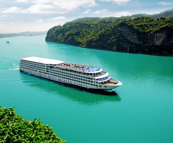 A large white cruise ship sails smoothly on a turquoise river, flanked by lush green hills. The sky is clear, conveying a serene and peaceful atmosphere.