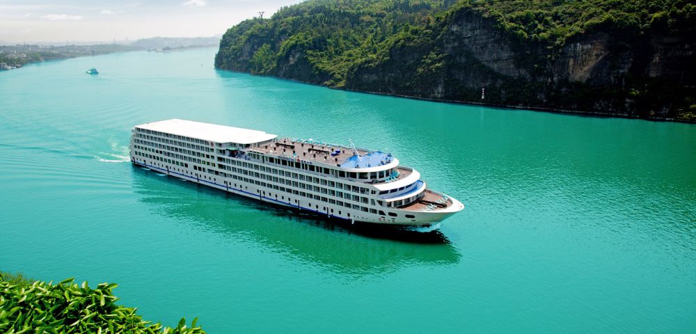 A large white cruise ship sails smoothly on a turquoise river, flanked by lush green hills. The sky is clear, conveying a serene and peaceful atmosphere.