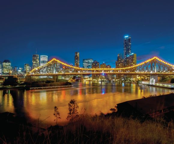 A brightly illuminated suspension bridge spans a calm river against a backdrop of a vibrant city skyline at night, reflecting lights on the water's surface.