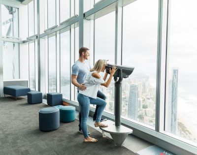A couple enjoys a panoramic city and ocean view from a skyscraper's observatory. The woman looks through binoculars as the man gently supports her.