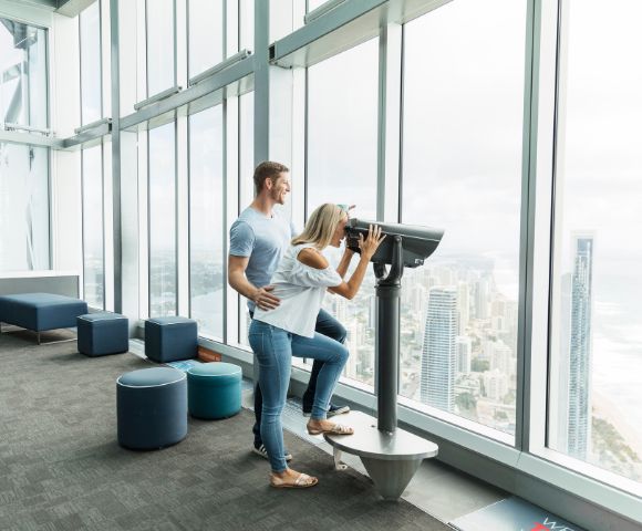 A couple enjoys a panoramic city and ocean view from a skyscraper's observatory. The woman looks through binoculars as the man gently supports her.