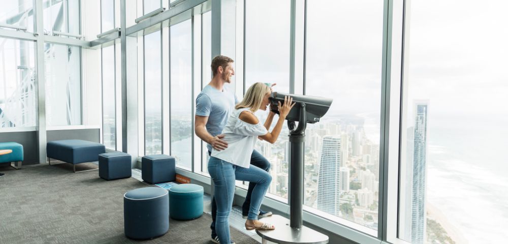 A couple enjoys a panoramic city and ocean view from a skyscraper's observatory. The woman looks through binoculars as the man gently supports her.