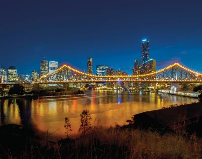 A brightly illuminated suspension bridge spans a calm river against a backdrop of a vibrant city skyline at night, reflecting lights on the water's surface.