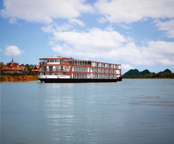 A large cruise ship sails on a calm river under a blue sky with scattered clouds. Lush green trees and distant hills line the riverbank, creating a serene and relaxing scene.