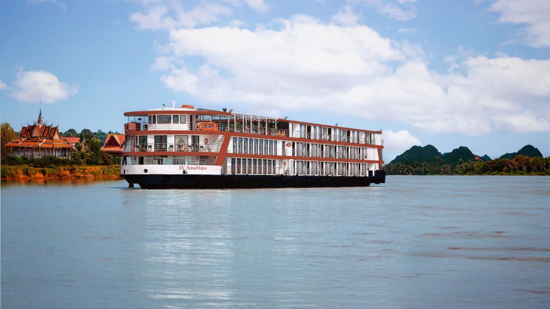 A large cruise ship sails on a calm river under a blue sky with scattered clouds. Lush green trees and distant hills line the riverbank, creating a serene and relaxing scene.