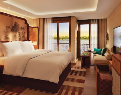 Luxurious hotel room with a large bed, elegant decor, and a private balcony overlooking a serene river. Cozy seating area and warm lighting create a tranquil ambiance.