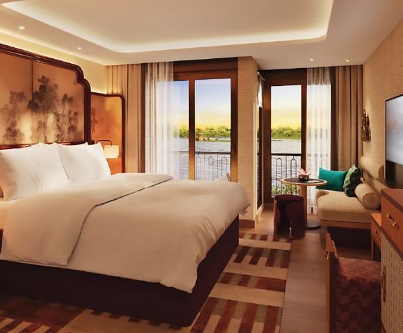 Luxurious hotel room with a large bed, elegant decor, and a private balcony overlooking a serene river. Cozy seating area and warm lighting create a tranquil ambiance.