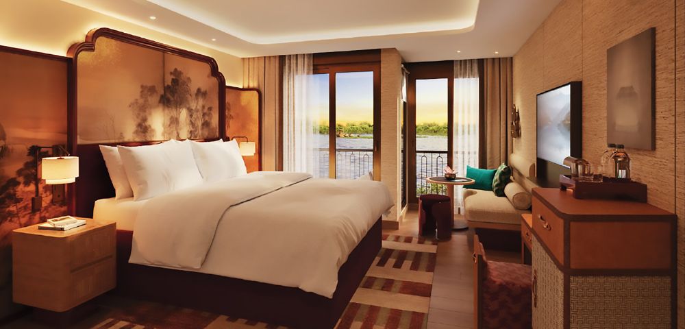 Luxurious hotel room with a large bed, elegant decor, and a private balcony overlooking a serene river. Cozy seating area and warm lighting create a tranquil ambiance.