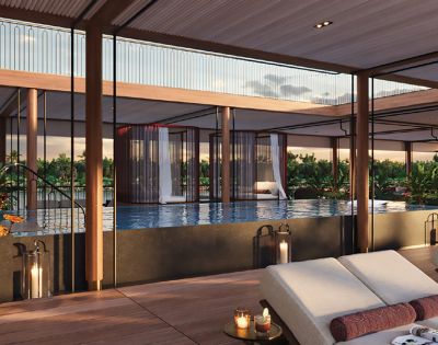 Luxurious poolside lounge featuring a modern deck, cushioned chairs, lit candles, and a serene infinity pool. Glass walls offer a sunset view of greenery.