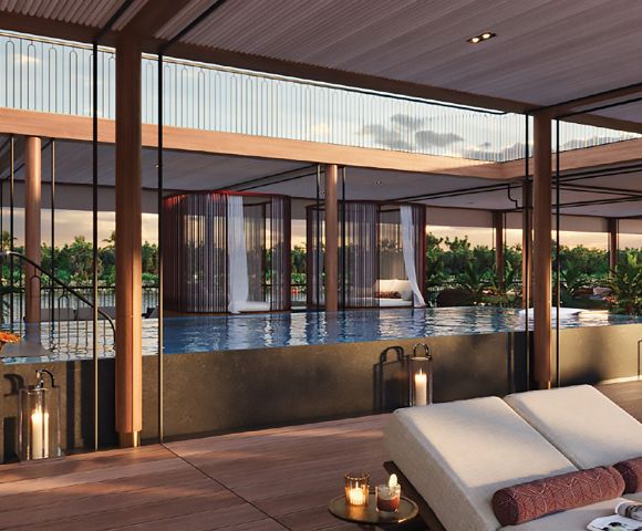 Luxurious poolside lounge featuring a modern deck, cushioned chairs, lit candles, and a serene infinity pool. Glass walls offer a sunset view of greenery.
