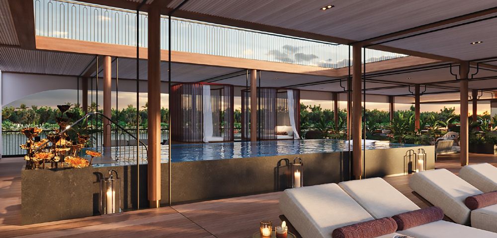Luxurious poolside lounge featuring a modern deck, cushioned chairs, lit candles, and a serene infinity pool. Glass walls offer a sunset view of greenery.