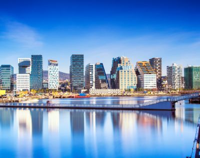 Skyline of a modern city with unique, colorful high-rise buildings reflected in calm water under a clear blue sky, conveying a serene atmosphere.
