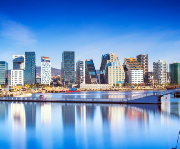 Skyline of a modern city with unique, colorful high-rise buildings reflected in calm water under a clear blue sky, conveying a serene atmosphere.