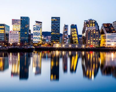 A city skyline at dusk, featuring modern, illuminated buildings reflected in calm water, creating a vibrant, mirrored effect. The atmosphere is tranquil.