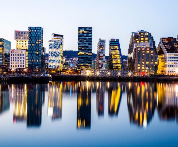 A city skyline at dusk, featuring modern, illuminated buildings reflected in calm water, creating a vibrant, mirrored effect. The atmosphere is tranquil.