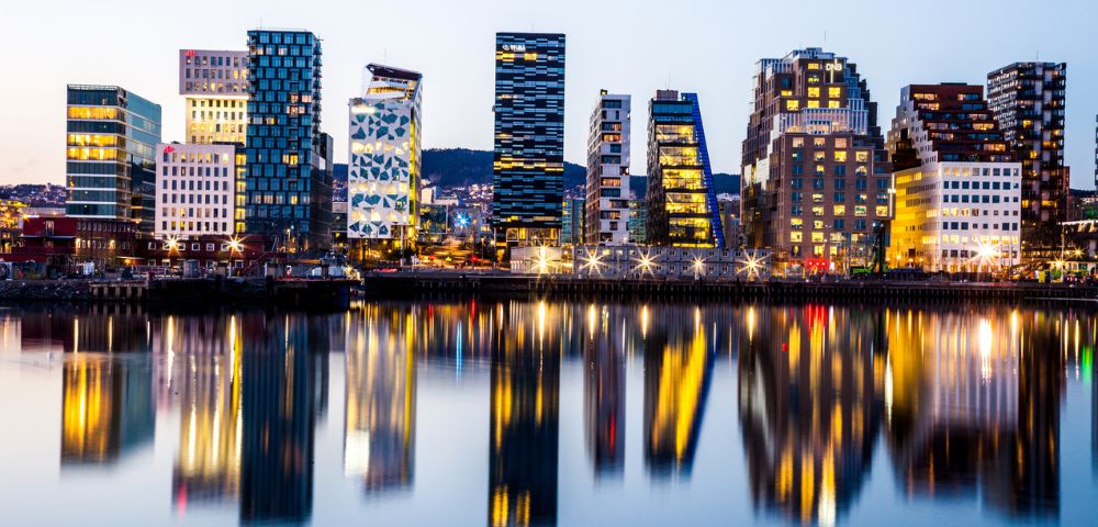 A city skyline at dusk, featuring modern, illuminated buildings reflected in calm water, creating a vibrant, mirrored effect. The atmosphere is tranquil.